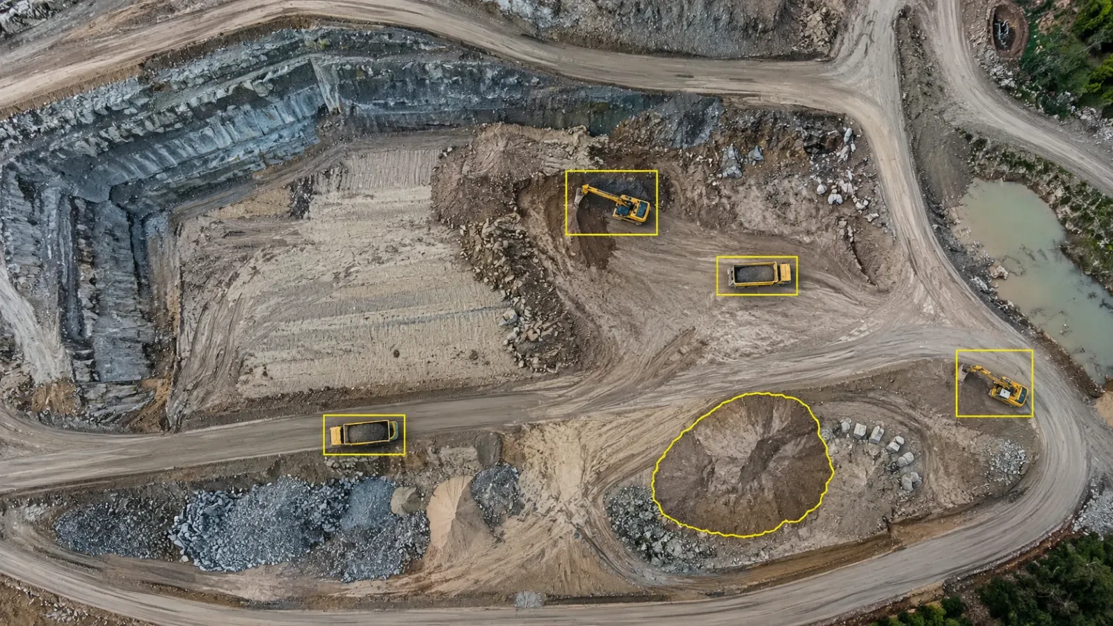 Top-down drone view of a large earthworks site under an overcast sky, with thin yellow boxes around a few excavators and dump trucks and one stockpile outlined