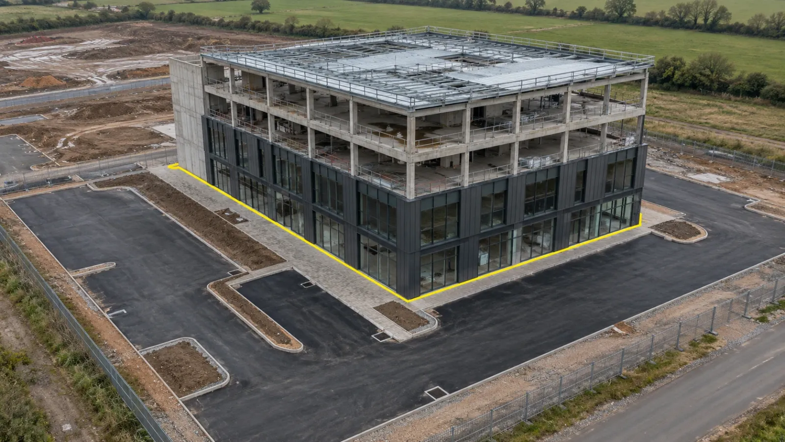 Oblique aerial view of a finished four-storey building frame on an overcast day, with new kerbs and paving around it and a thin yellow outline tracing the building footprint