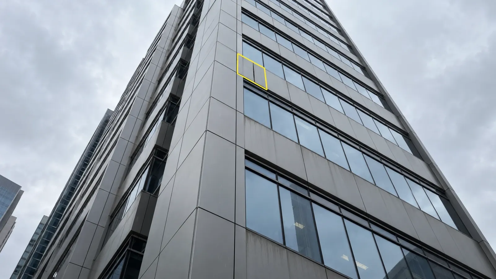 Commercial office tower clad in grey metal panels and glazing seen from street level under an overcast sky, with one cladding panel high on the wall outlined in yellow