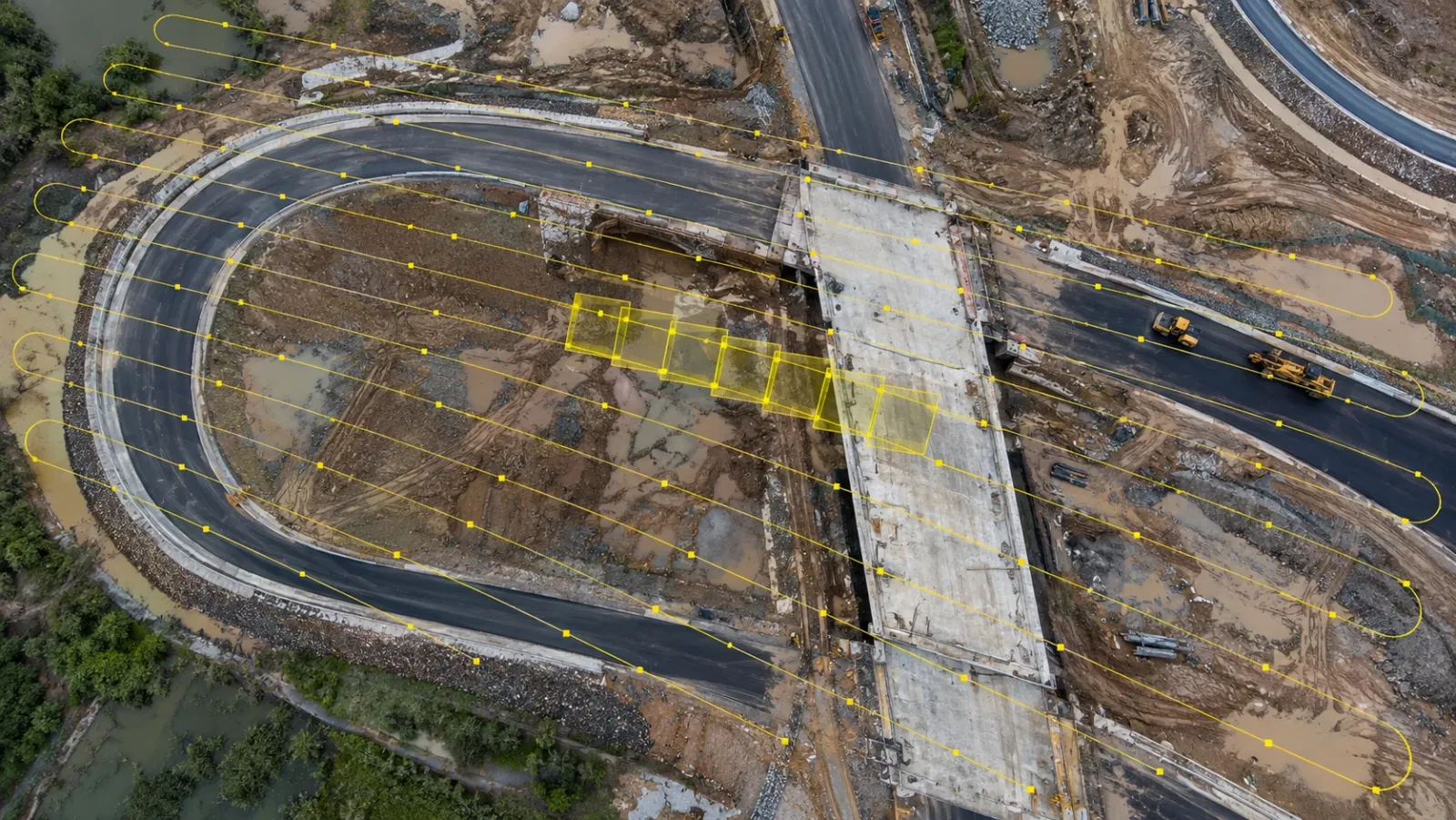 Top-down view of a road interchange under construction after rain, with a yellow lawnmower flight path and overlapping photo footprints drawn across it