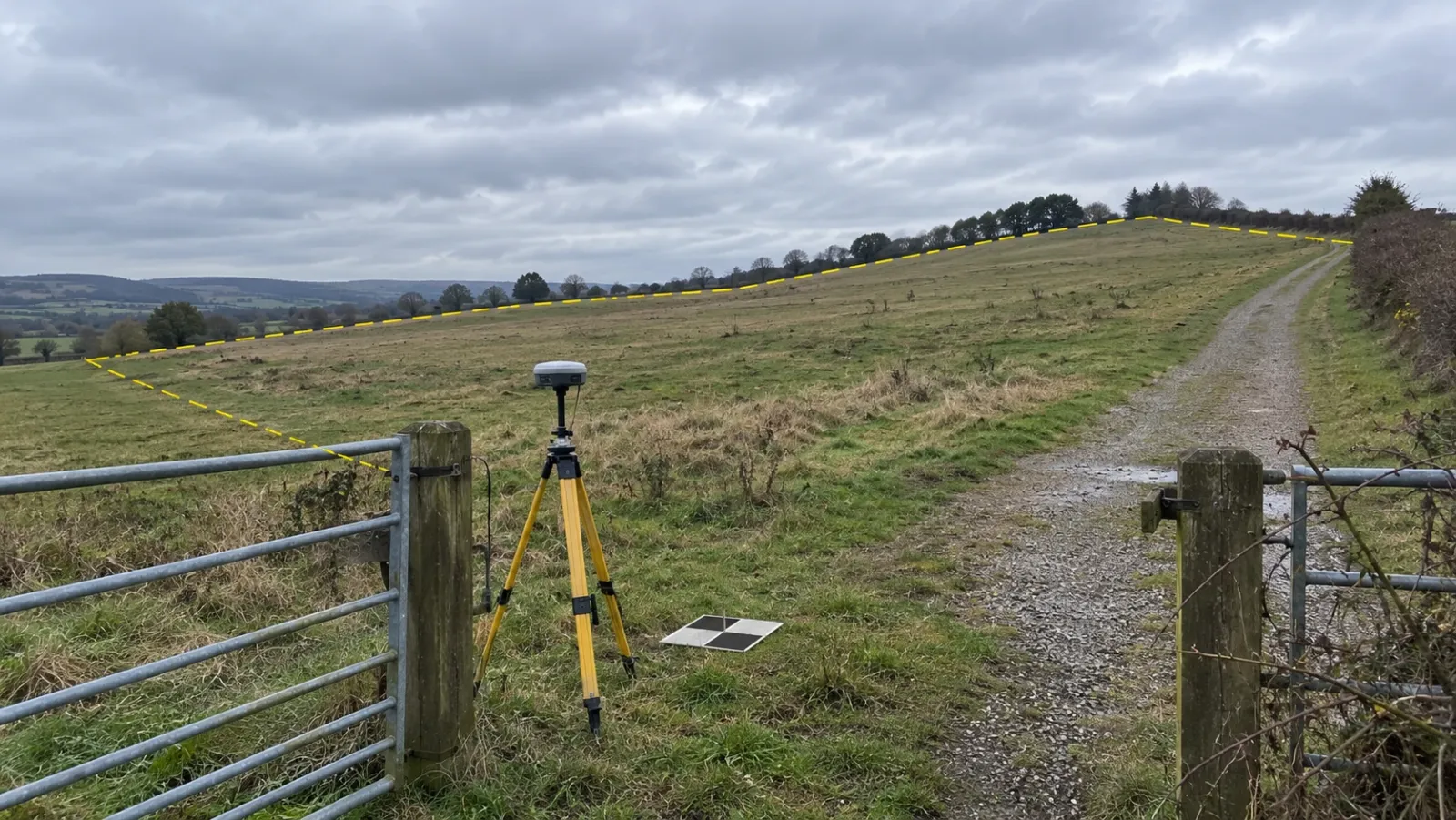 Undeveloped sloping parcel seen from a field gate under an overcast sky, with a survey tripod and a ground control target in the grass and a thin yellow line tracing the parcel boundary