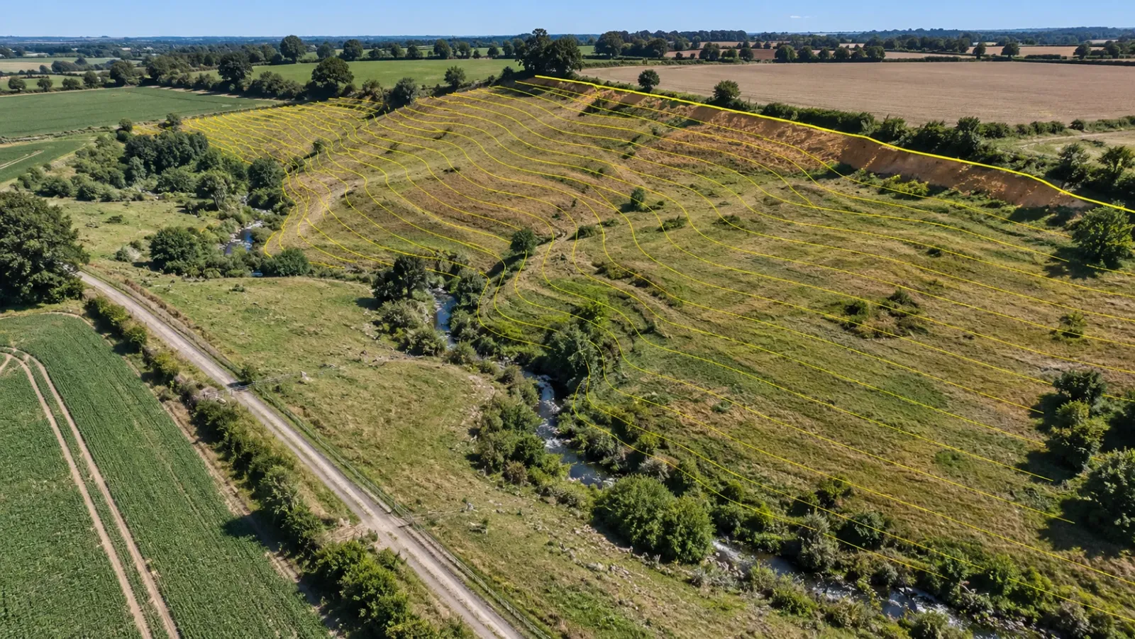 High oblique aerial view of a sloping undeveloped parcel at midday with a stream gully, a farm track and yellow contour lines bending into the gully