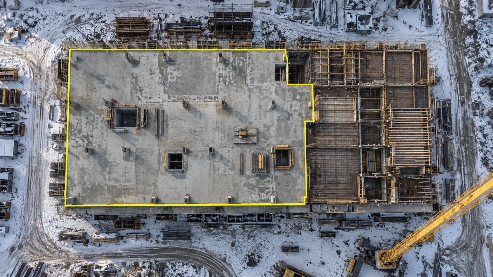 Top-down drone view of a concrete building frame on a snowy winter morning, with a thin yellow outline around only the slab bays that are poured and stripped