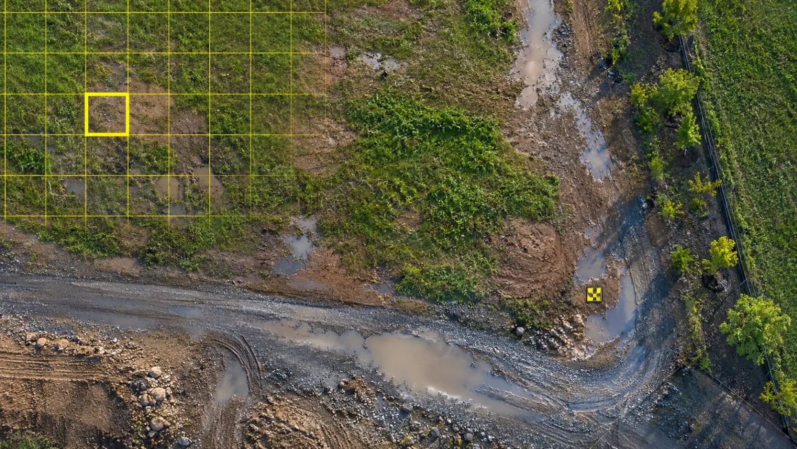 Top-down drone view of an open development parcel just after rain, with puddles reflecting the sky and a fine yellow pixel grid over one corner