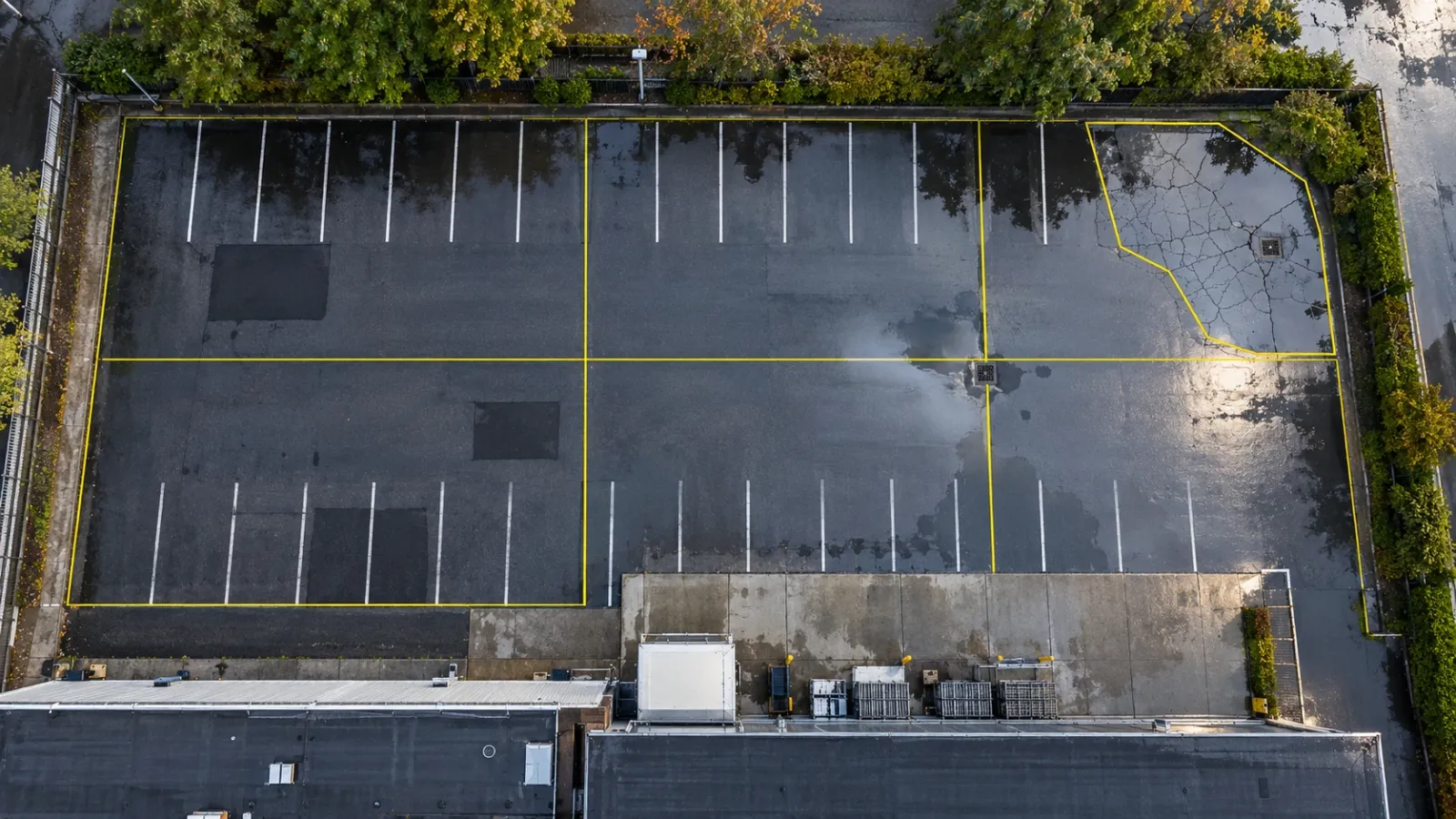 Top-down drone view of an empty asphalt car park and service yard after rain, with puddles in low spots, patched areas and thin yellow lines dividing the paving into survey sections