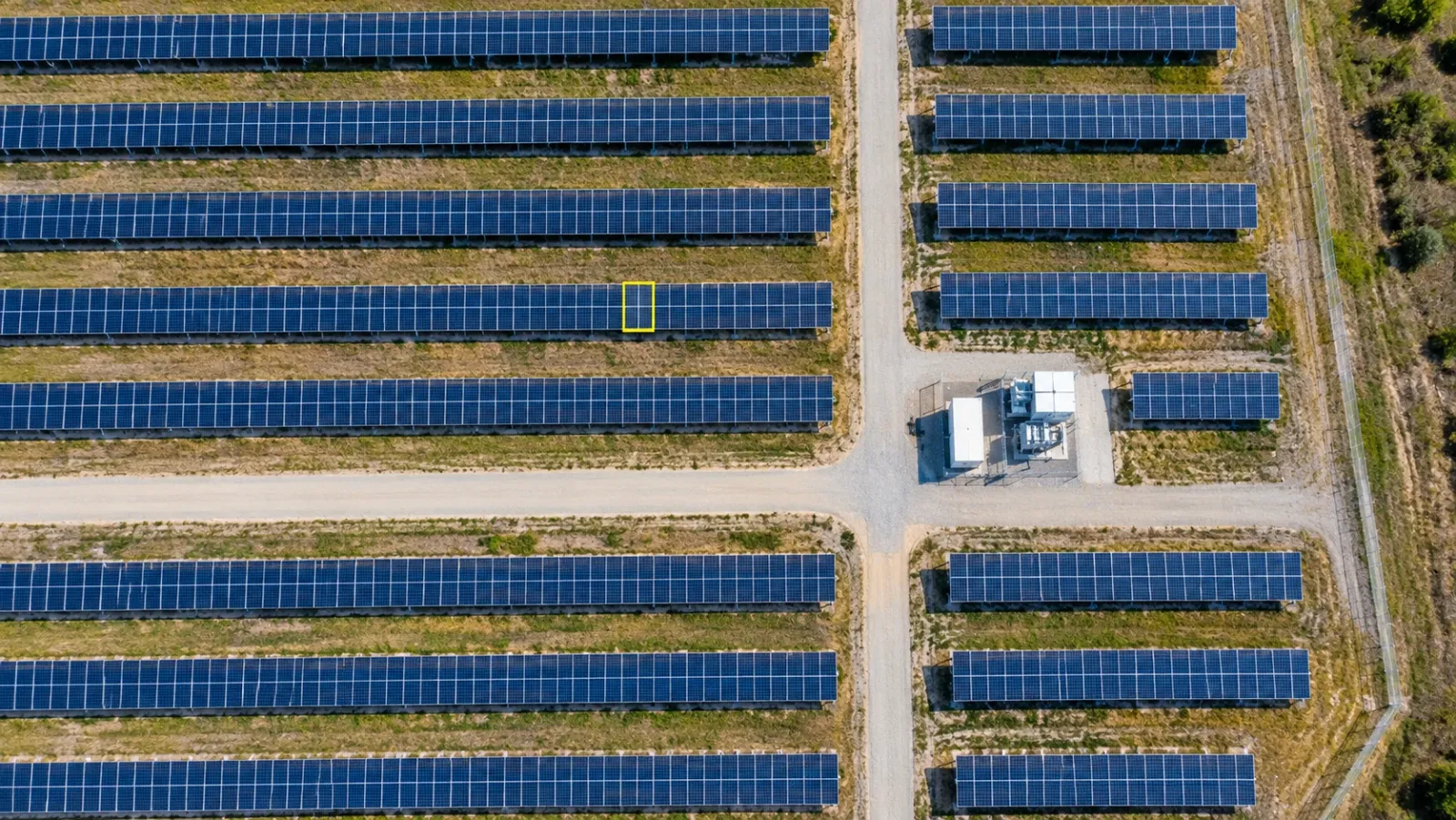 Top-down aerial view of a utility-scale solar farm at midday, rows of panels split by gravel tracks around an inverter station, with one module outlined in yellow