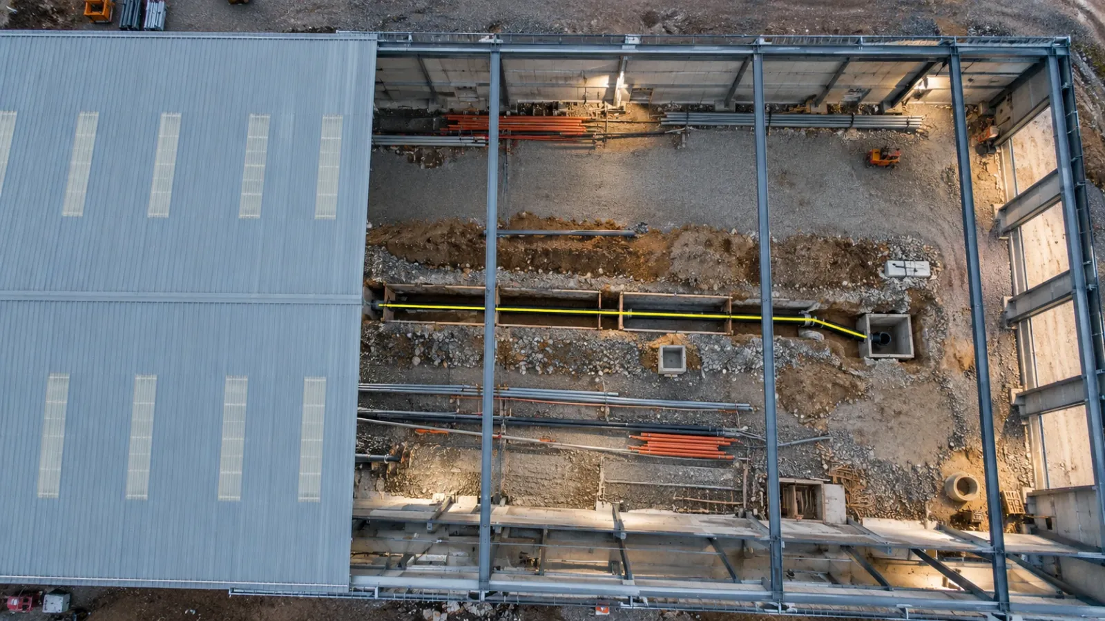 Top-down view through the open roof bays of a warehouse shell into a service trench lit by work lights, with ducts and a drainage pipe on a sand bed and a thin yellow line along the pipe