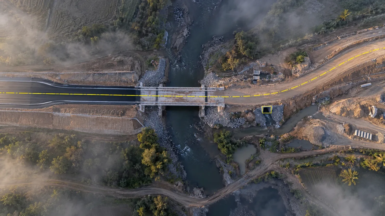 Top-down drone view of a new road and bridge under construction in early morning mist, with a thin yellow design centreline traced along the carriageway and one culvert marked