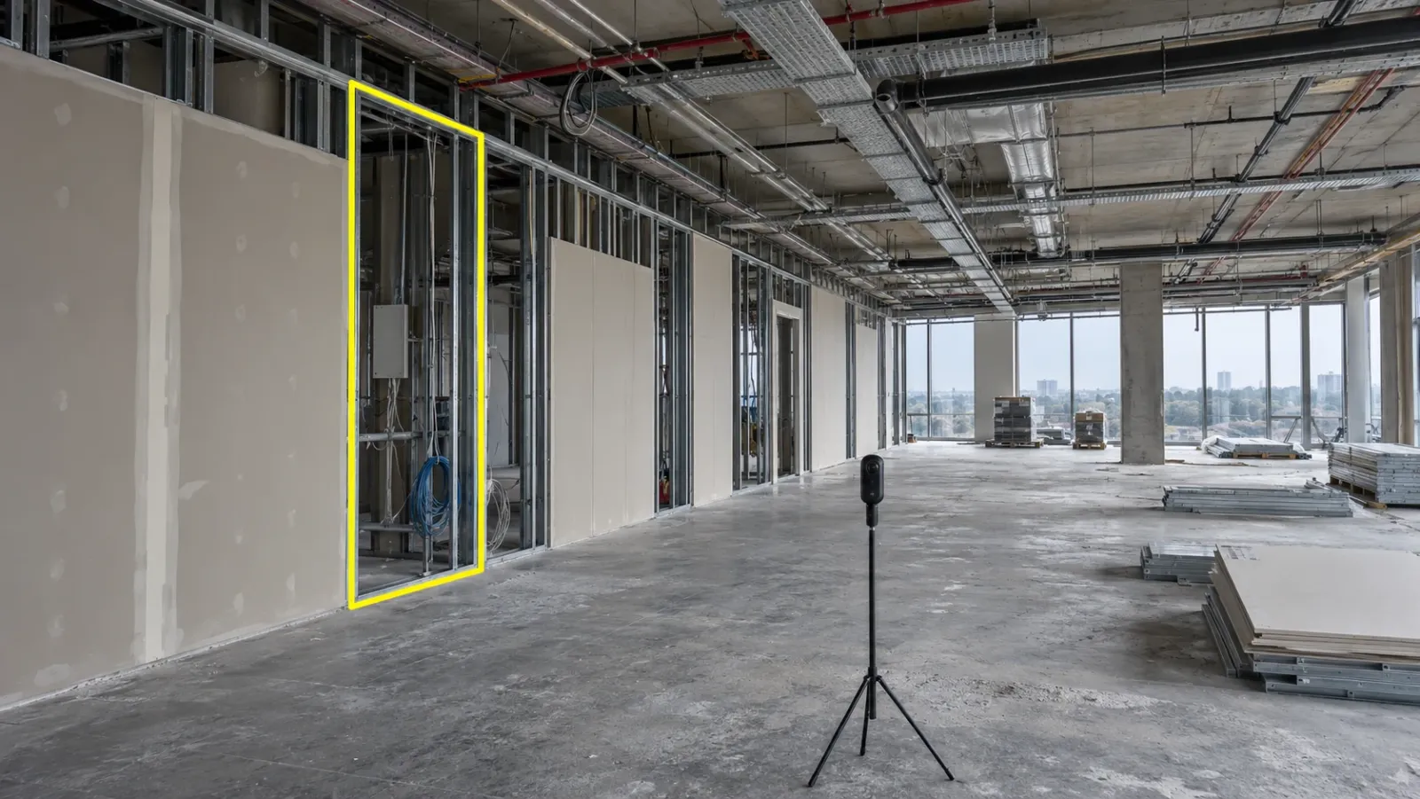 Wide interior view of an office floor mid fit-out in soft overcast daylight, with a 360° camera on a tripod and one unfinished partition bay outlined in yellow